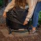 A woman places a protective cover over a TIKI® Brand fire pit on a patio, surrounded by outdoor greenery.