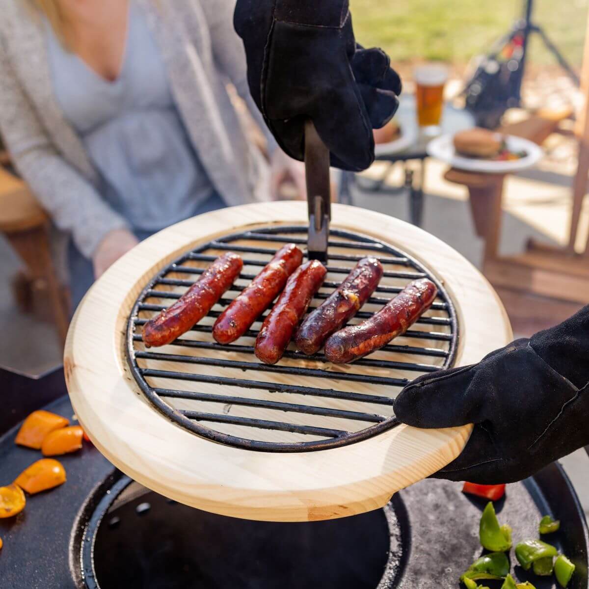 Gloved hands lift a grill grate with cooked sausages from a TIKI fire pit; outdoor gathering in background.