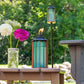 TIKI Brand turquoise table torch with flame, next to a mason jar of flowers on an outdoor patio, green trees in background.