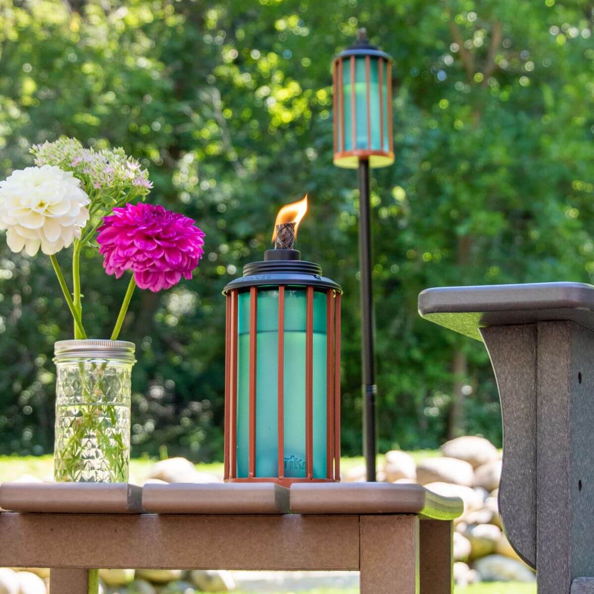 TIKI Brand turquoise table torch with flame, next to a mason jar of flowers on an outdoor patio, green trees in background.