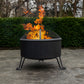 Black outdoor fire pit with flames, on concrete, near green hedge.