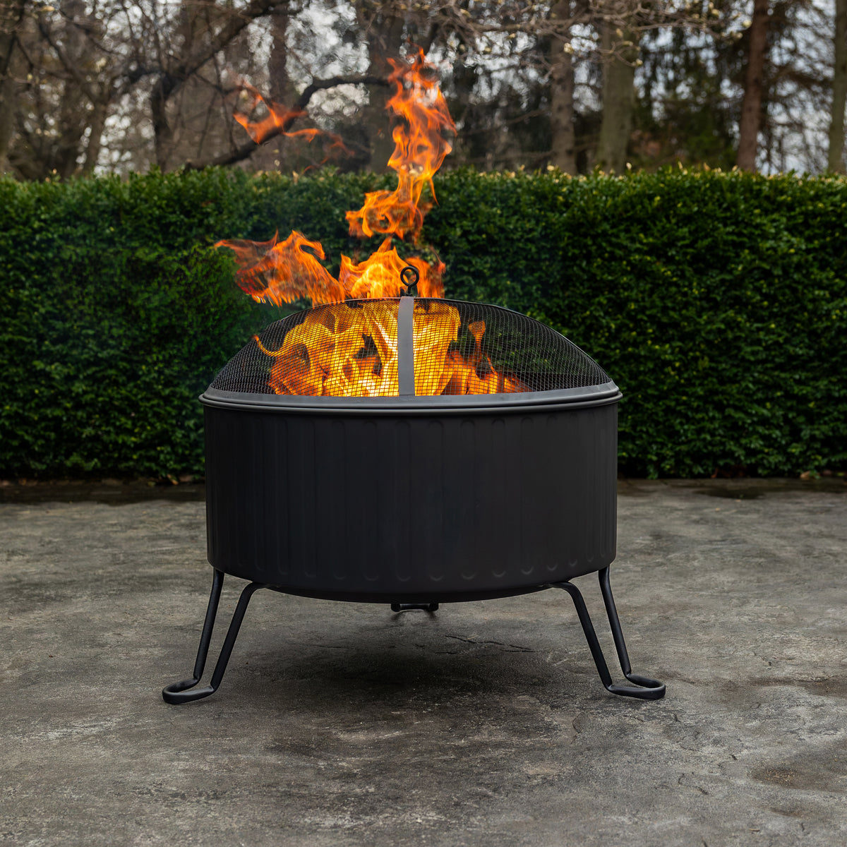 Black outdoor fire pit with flames, on concrete, near green hedge.